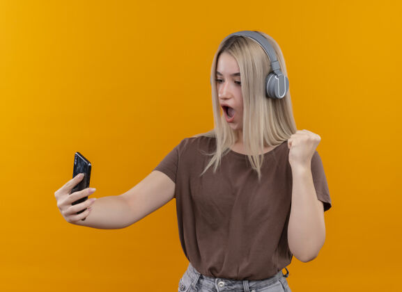 拳头惊讶的年轻金发女孩戴着耳机拿着手机在孤立的橙色墙上举起拳头看着它耳机提高年轻