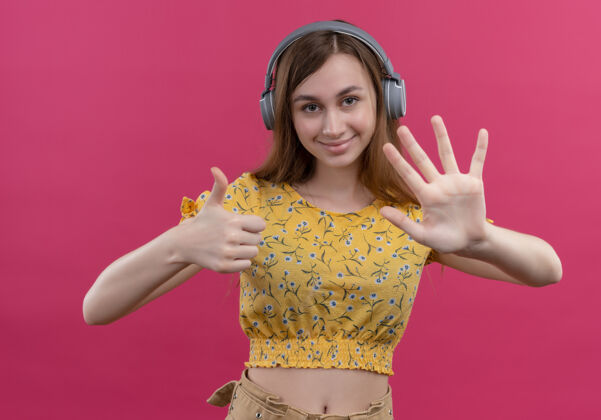 年轻微笑着戴着耳机的年轻女孩 在孤立的粉色墙壁上展示着五只手和拇指耳机女孩微笑