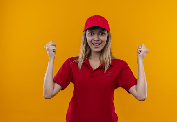 微笑微笑着的年轻送货女孩穿着红色制服 戴着帽子 在橙色的墙上显示“是”的手势年轻女孩显示