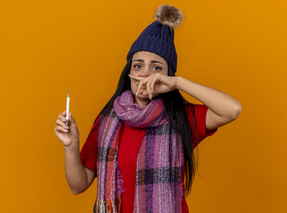 疾病瘦弱的年轻白种人生病的女孩戴着冬天的帽子和围巾拿着温度计擦鼻子隔离在橙色的墙上穿体温计帽子