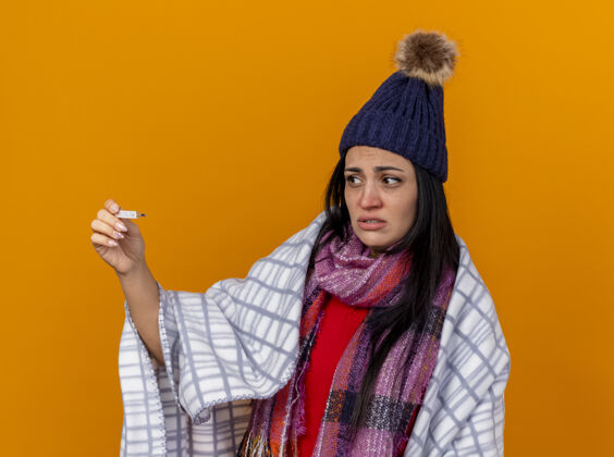围巾焦急的年轻白种人生病的女孩戴着冬天的帽子 围着格子呢围巾 手里拿着隔离在橙色墙上的温度计穿帽子格子