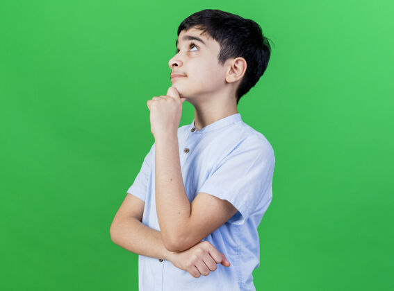 侧写体贴的年轻白人男孩站在侧视图触摸下巴仰望隔离在绿色墙上的复制空间下巴站立年轻