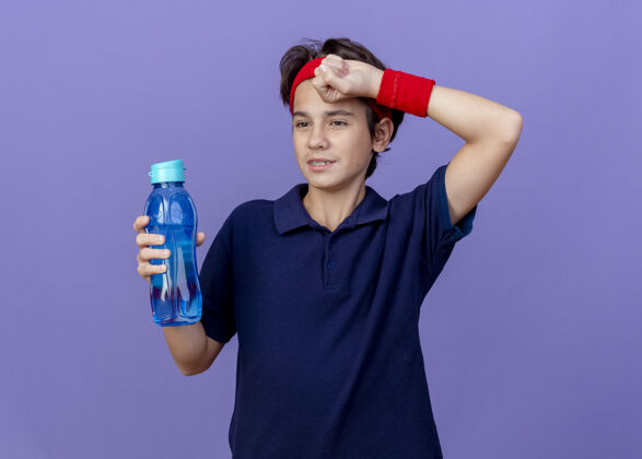 湿巾疲惫的年轻帅气的运动男孩戴着头带和护腕戴着牙套看着一边拿着水瓶擦着汗水用手隔离在紫色的墙上瓶子男孩疲倦