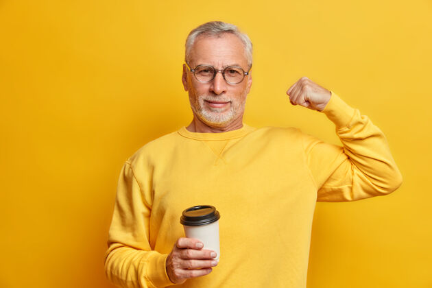 情绪严肃自信的成熟男人举起手臂 露出强壮有力的肱二头肌拿着咖啡纸杯穿着休闲套头衫隔着黄色的墙壁男人年龄成熟