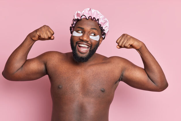 治疗快乐坚强微笑的男人举起双臂展示肱二头肌赤裸地站在室内靠着粉色墙壁进行整容手术戴上润肤贴在眼睛下面防水浴帽人人产品