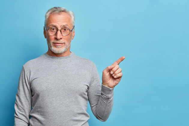 胡须好奇的白发老人用食指指着空白处的水平镜头显示出你的广告位置穿着休闲套头衫隔着蓝色的墙壁人指向自信
