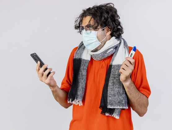 男人印象深刻的年轻白种人病人戴着眼镜 围巾和口罩拿着温度计和手机看着隔离在白墙上的手机手机年轻人印象