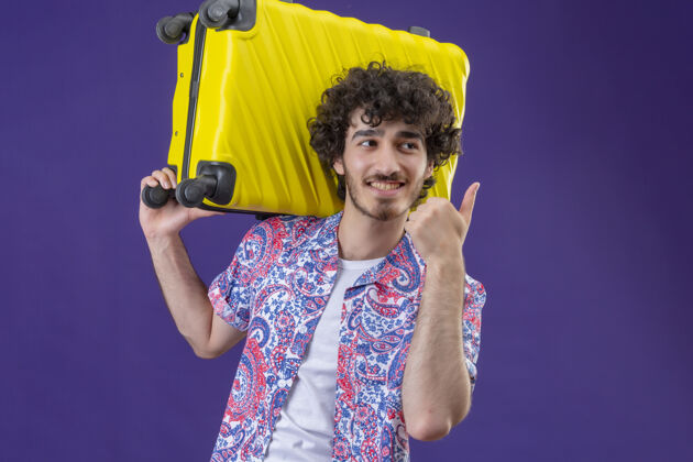 卷曲微笑的年轻英俊的卷发旅行者男子肩上提着手提箱 竖起大拇指 看着右边的紫色隔离墙 上面留着复制空间微笑肩膀右