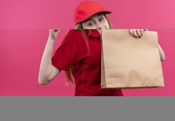 女孩微笑着的年轻送货女孩 穿着红色制服 握紧拳头拿着纸袋 站在孤立的粉色墙上年轻包举行
