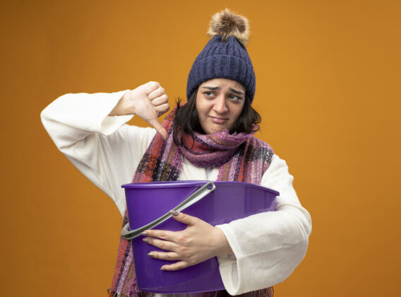 年轻年轻的高加索病女孩穿着长袍 冬天帽子和围巾有恶心拿着塑料桶看一边显示拇指向下孤立在橙色的墙上与复制空间穿冬天塑料