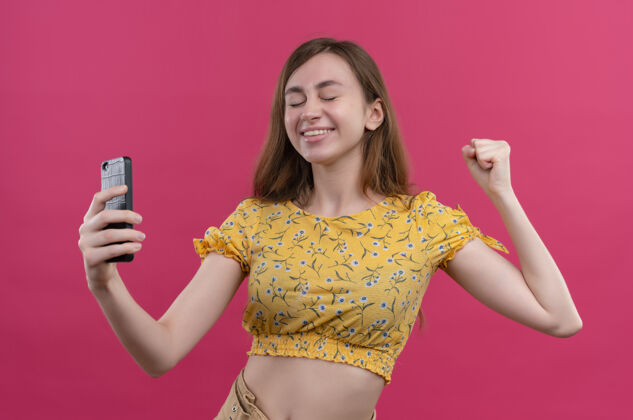 手机微笑的年轻女孩拿着手机 闭着眼睛举起拳头对着孤立的粉色墙壁握着举起封闭