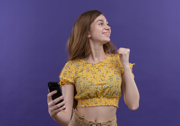 手机微笑的年轻女孩拿着手机 举起拳头看着孤立的紫色墙上的右侧右举起微笑
