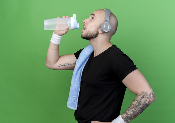 年轻人站在侧视图中 戴着护腕和耳机的年轻运动型男子从水瓶里喝水 肩膀上的毛巾被隔离在绿色的墙上瓶子肩膀立场