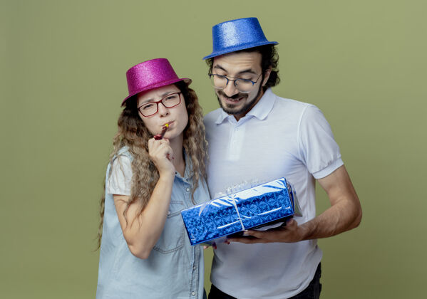 吹口哨一对年轻夫妇戴着粉色和蓝色的帽子 女孩吹着口哨 男孩拿着礼物盒看着橄榄绿的墙上孤立的礼物盒奥利弗情侣小伙子