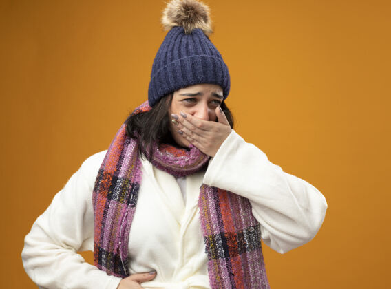 长袍疼痛的年轻白种人生病的女孩穿着长袍 戴着冬天的帽子和围巾恶心把手放在肚子上和嘴上看着橙色墙上孤立的一面年轻女孩疾病