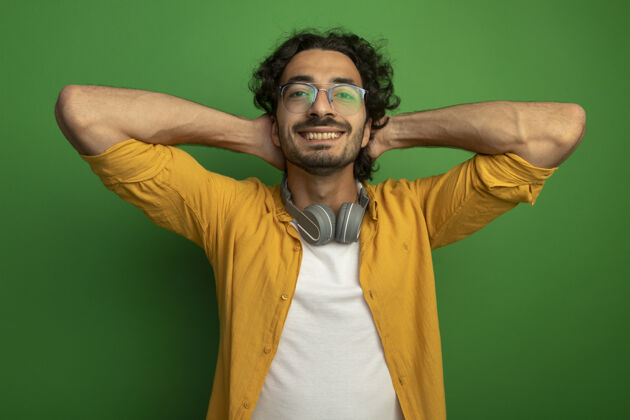 围着微笑着的年轻英俊的白人男子戴着眼镜 脖子上戴着耳机 手放在头后 隔离在绿色的墙上微笑耳机保持