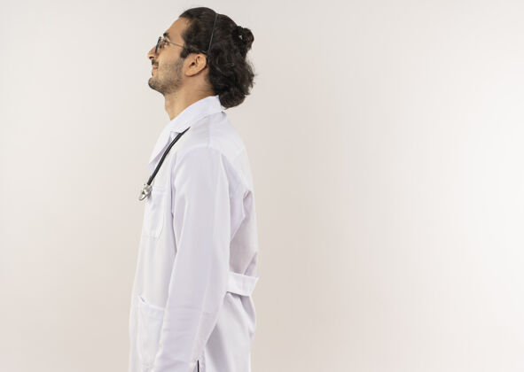 医生站在剖面图上的年轻男性医生戴着眼镜 穿着白色长袍 听诊器放在隔离的白色墙壁上 留有复印空间长袍听诊器个人资料