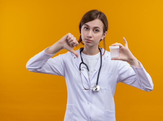 年轻年轻的女医生穿着医用长袍 手持听诊器 拿着烧杯 拇指朝下 站在孤立的橙色墙壁上烧杯女人不愉快
