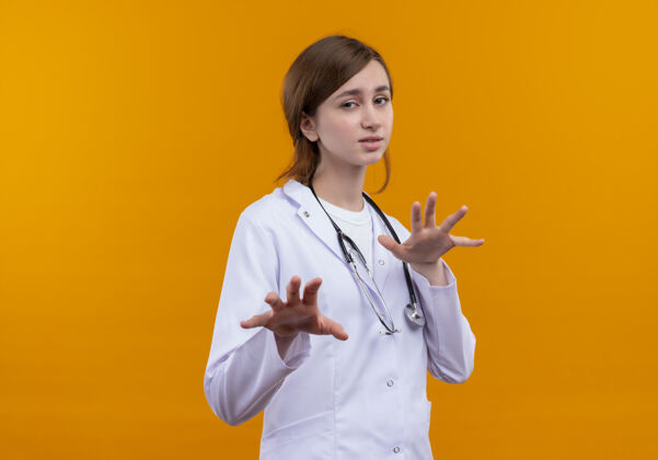 医生穿着医用长袍和听诊器的年轻女医生在隔离的橙色墙壁上伸展双手 并留有复印空间不愉快听诊器穿