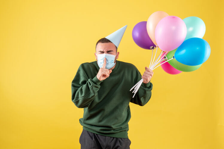 大流行正面图：黄色背景上 年轻男性手持彩色气球 戴着无菌面具消毒年轻男性庆祝