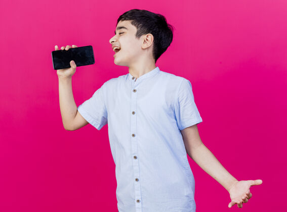 电话年轻的高加索男孩唱歌 用手机作为麦克风 在深红的背景下显示出空手男孩麦克风手机
