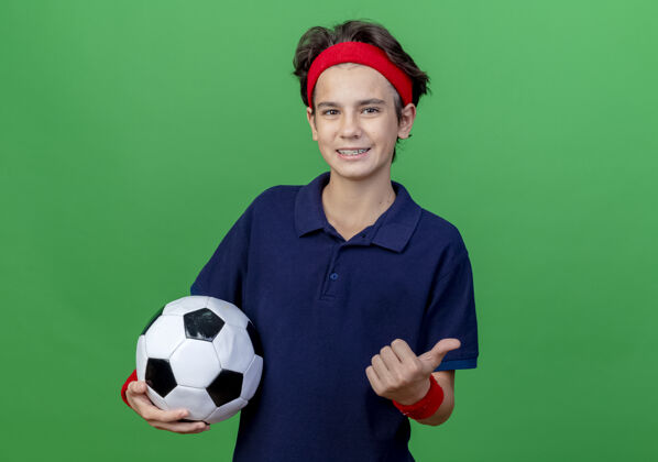 男孩微笑的年轻帅气的运动男孩戴着头带和护腕 戴着牙套 举着足球 拇指朝上 看着隔离在绿色背景上的相机 还有复制空间拇指运动牙套