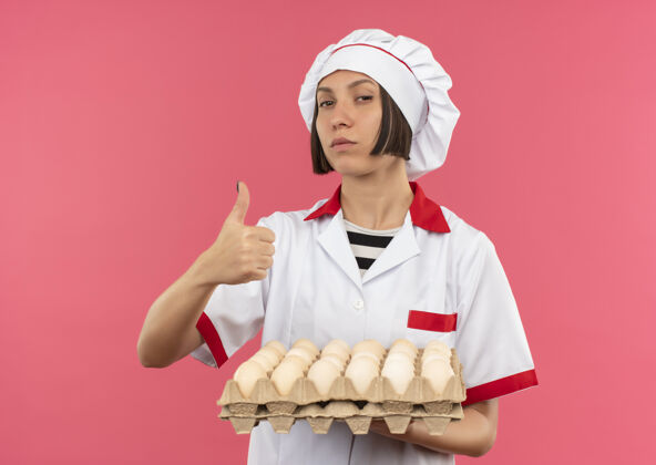 制服自信的年轻女厨师身着厨师制服 手里拿着一盒鸡蛋 在粉红色的地板上孤立地竖起大拇指厨师年轻纸箱