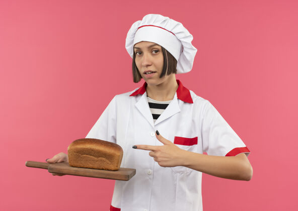 年轻自信的年轻女厨师穿着厨师制服 拿着并指着切菜板 上面的面包是粉红色的厨师制服面包