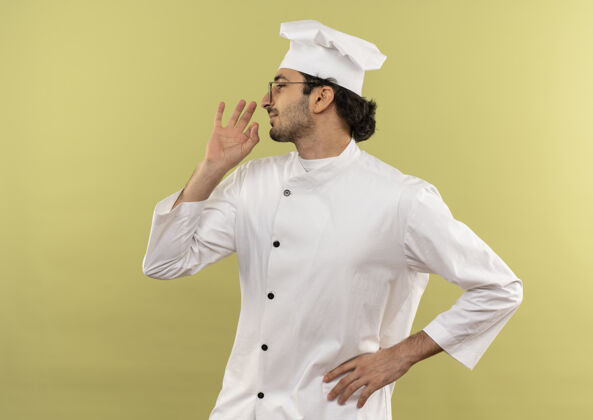 厨师站在侧视图中 年轻的男厨师穿着厨师制服 戴着眼镜 展示着美味的手势 手放在绿色墙壁上的臀部显示制服眼镜
