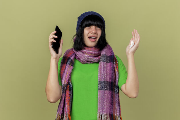 印象印象深刻的年轻病高加索女孩戴着冬天的帽子和围巾手持手机和餐巾看着一边举起手在橄榄绿的背景与复制空间隔离手机壁板女孩