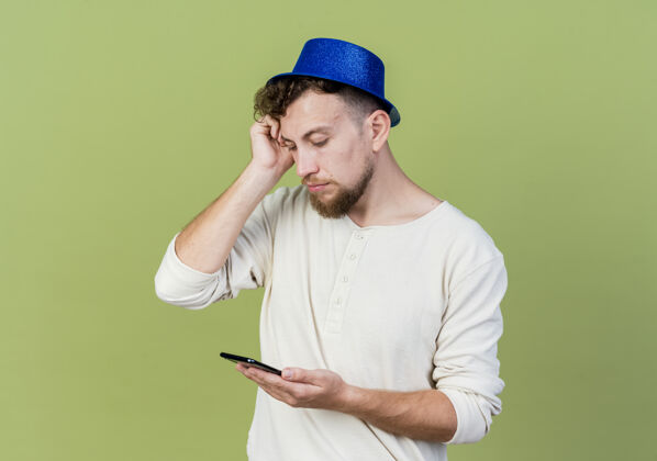 年轻体贴的年轻帅哥斯拉夫党的家伙戴着党的帽子举行 并看着手机触摸头孤立在橄榄绿的背景与复制空间头小伙子手机