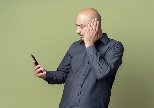 手持印象深刻的年轻秃头呼叫中心男子手持手机 看着与橄榄绿孤立的头上的手秃头头电话
