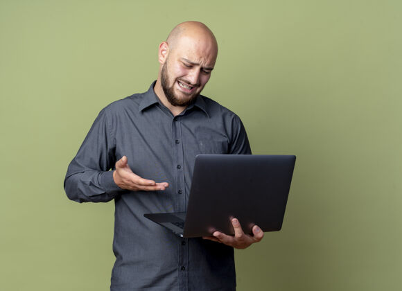 指向恼怒的年轻秃头呼叫中心男子拿着和看笔记本电脑 并用手指着它孤立的橄榄绿与复印空间年轻电话手