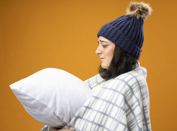 虚弱体弱多病的白种年轻女孩 穿着长袍 戴着格子呢的冬帽 站在侧视图中 拥抱着 看着孤立在橙色背景上的枕头疾病格子包裹