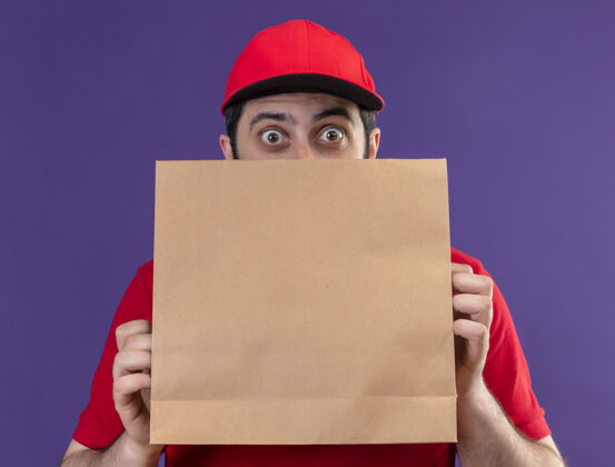送印象深刻的年轻英俊的白人送货员身穿红色制服 戴着帽子 从后面看 纸包裹上孤立的紫色男帽子穿着