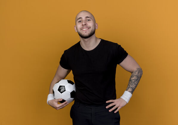 微笑微笑的年轻运动男子戴着手环拿着球 把手放在臀部隔离在橙色的墙上持有运动年轻