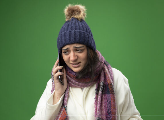 围巾穿着长袍 戴着冬帽 戴着围巾的年轻白种人生病的女孩在打电话 看着绿色背景下孤立的一面女孩电话不愉快