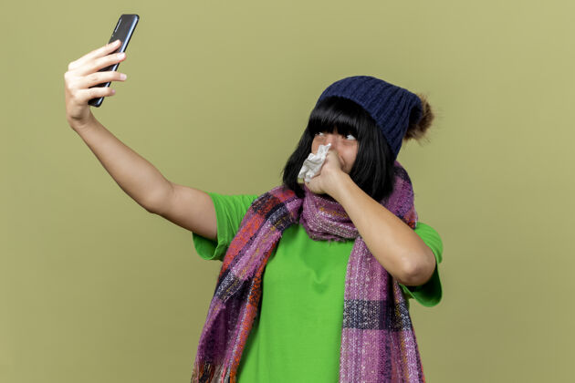 自拍年轻生病的高加索女孩戴着冬天的帽子和围巾自拍拿着餐巾手放在嘴上橄榄绿的背景与复制空间隔离采取餐巾手