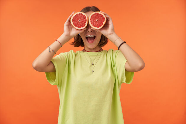 时髦女学生 一位年轻的女士 深色短发 眼睛上蒙着葡萄柚神情惊讶站在橙色的墙上穿着绿色t恤 戴着牙套和手镯20多岁背带热带