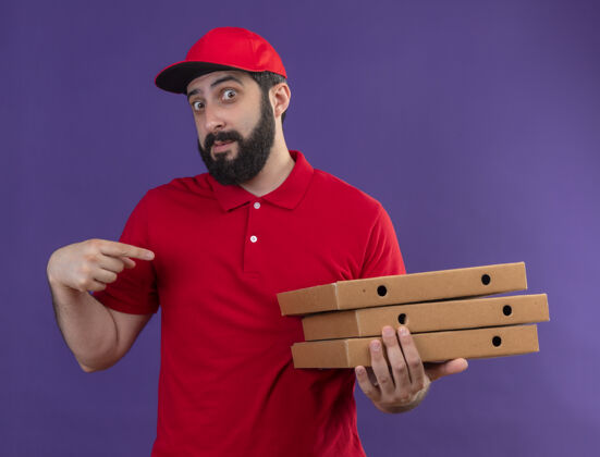 男人年轻帅气的白人送货员穿着红色制服 戴着帽子 指着紫色的比萨饼盒紫色帅气指着
