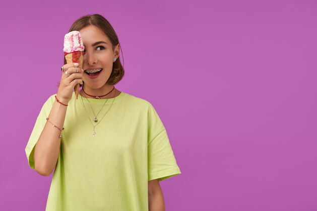 女士十几岁的女孩 开朗快乐 深色短发拿着冰激凌遮住眼睛 看着紫色墙上的复制空间穿着绿色t恤 戒指和项链右穿孔成人