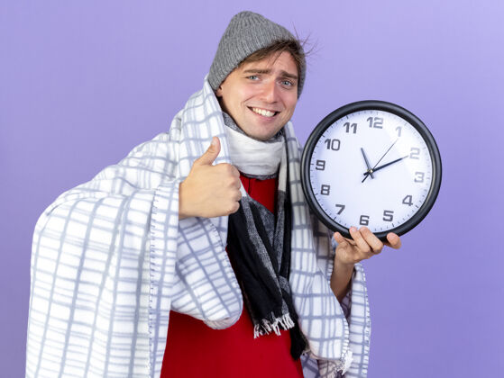 帽子微笑的年轻英俊的金发病患戴着冬天的帽子和围巾包裹在格子布拿着时钟看着相机显示拇指向上孤立的紫色背景时钟年轻围巾