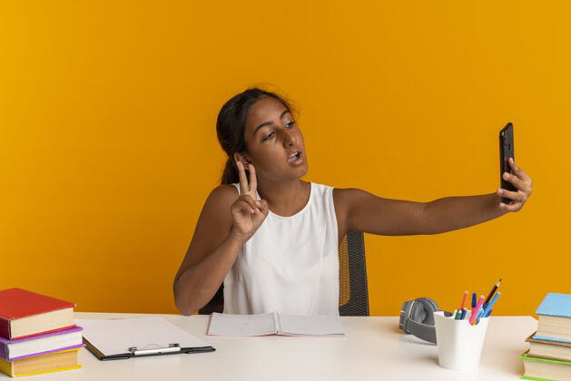 采取坐在书桌旁 拿着学习用具的小女孩高兴地自拍 在橘色的墙上表现出一种与世隔绝的和平姿态女学生工具书桌