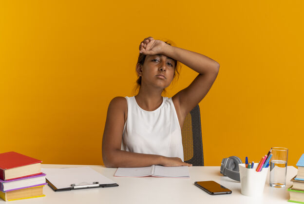 累疲惫的年轻女学生坐在书桌旁 把手腕放在额上 孤立地贴在橘色的墙上书桌学校工具