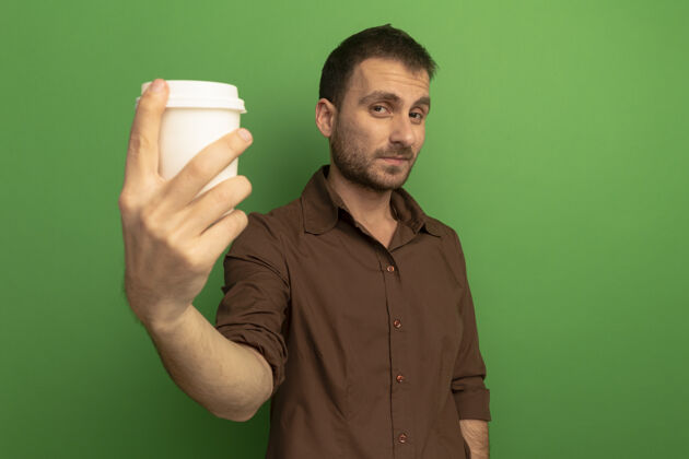 伸展年轻的白种人高兴地伸出塑料咖啡杯对着镜头 看着隔离在绿色背景上的相机 还有复印空间咖啡朝向请
