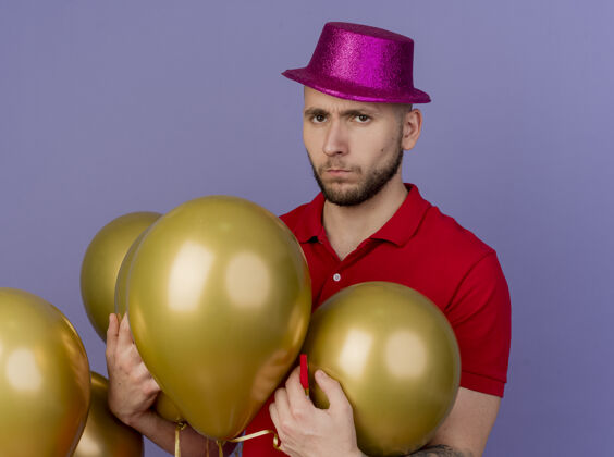 派对年轻帅气的斯拉夫派对男子戴着派对帽站在气球旁抓着气球 看着紫色背景上孤立的摄像机气球帅哥小伙子