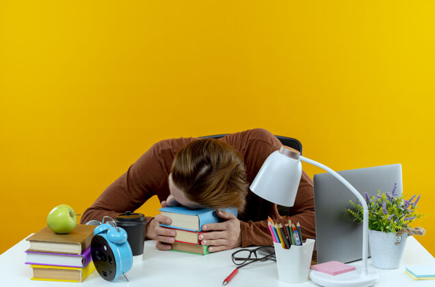 学校疲倦的年轻学生男孩坐在书桌旁 手里拿着学习工具 把头放在隔离在黄色墙上的书上年轻工具头