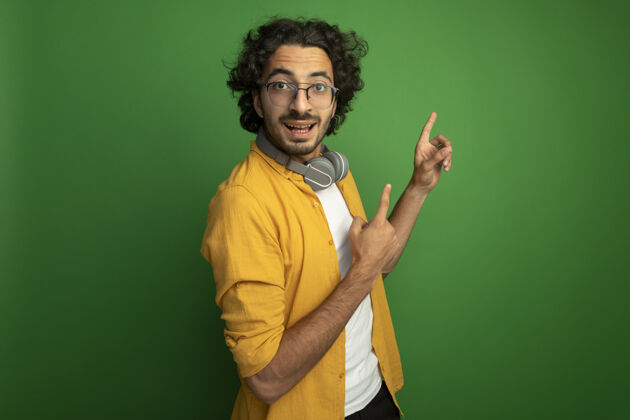 年轻印象深刻的年轻英俊的白人男子戴着眼镜戴着耳机站在侧视图看相机指向上孤立的绿色背景与复制空间相机围着眼镜