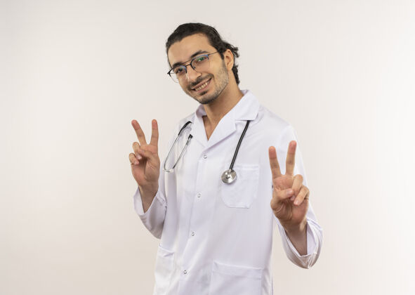光学微笑的年轻男医生 戴着眼镜 穿着白色长袍 带听诊器 在隔离的白色墙壁上展示着和平的姿态 还有复印空间听诊器长袍和平