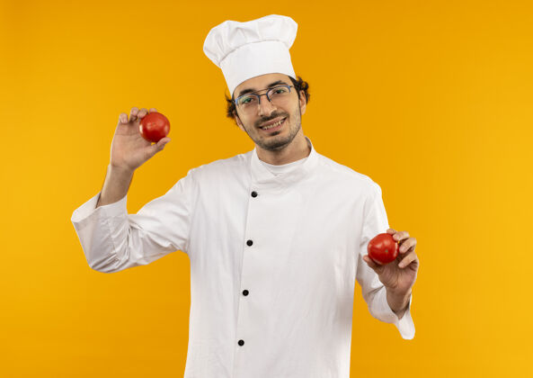 微笑面带微笑的年轻男厨师穿着厨师制服 戴着眼镜 手里拿着隔离在黄色墙上的西红柿烹饪男性穿着